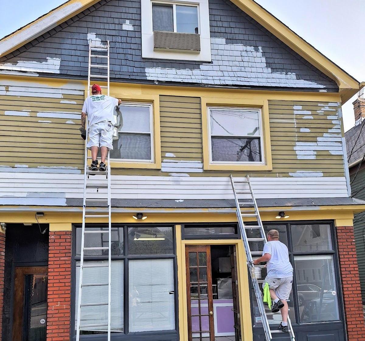 The front of a two-story building mid-job: gray-blue gable and olive siding scraped down to bare patches, with a crew member on a ladder and a brick storefront below.