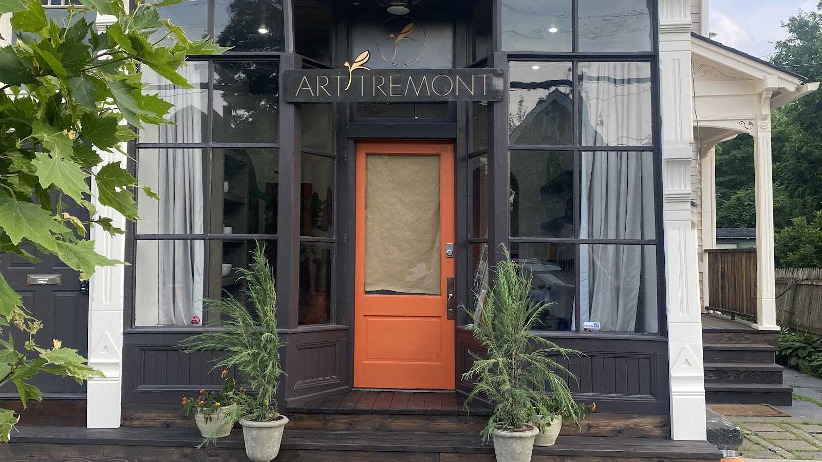 A dark-trimmed storefront with a bright orange door, a hanging sign above it and two potted evergreens on the front step.