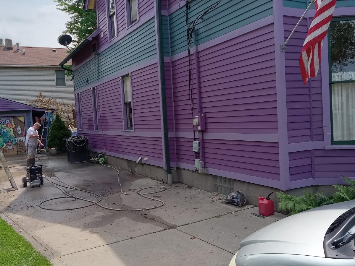 The side of a purple house with teal panels and lilac trim, a man power washing the driveway and an American flag on the porch.